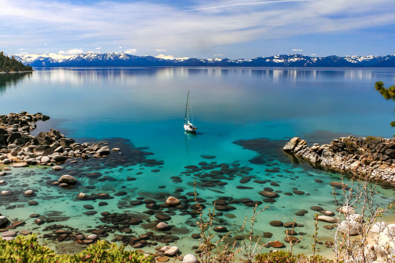 Tranquil blue waters of a lake with a solitary sailboat, surrounded by smooth pebbles and a backdrop of snow-capped mountains.
